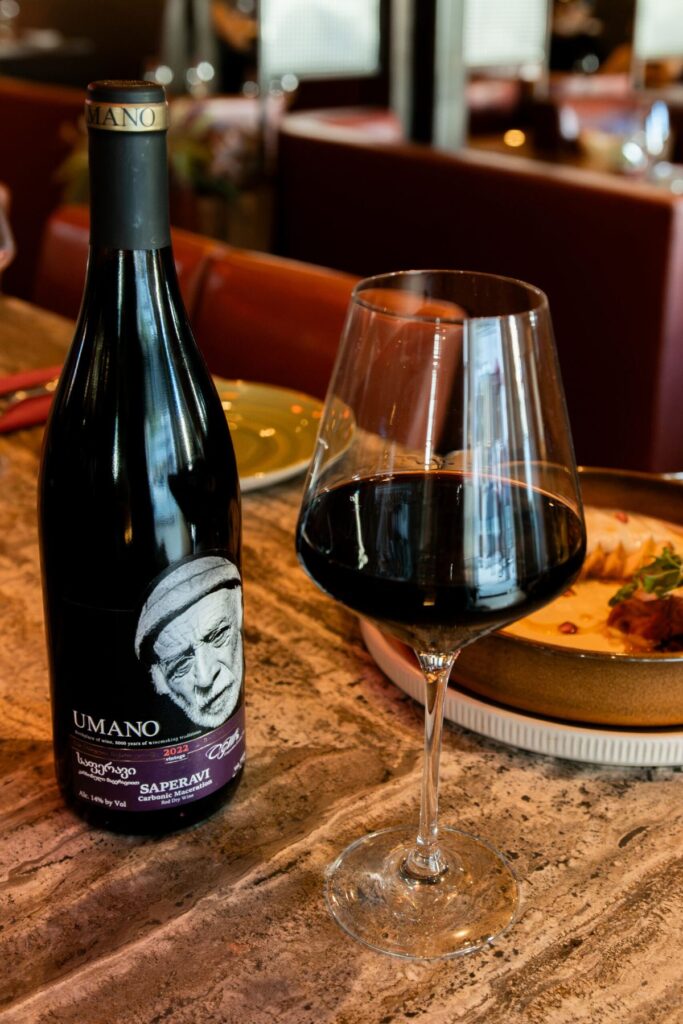 A bottle of red wine and a glass of red wine on a table beside a plated dish